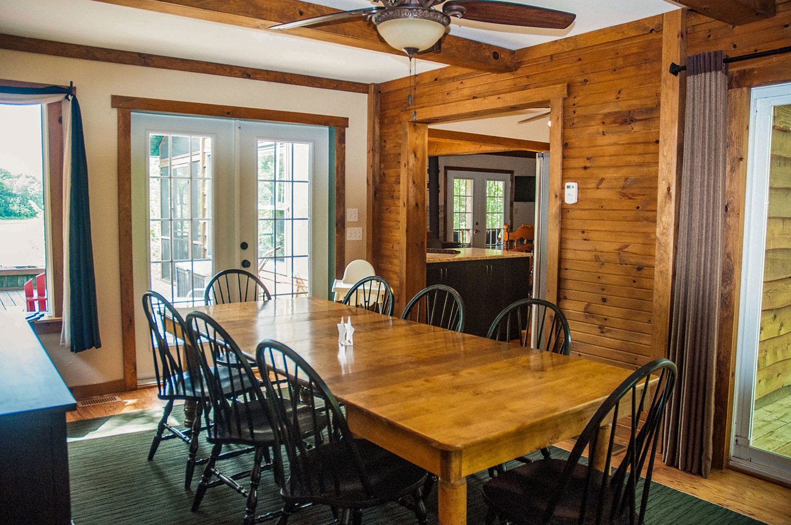 A spacious dining area with a large wooden table comfortably seats up to 10 people. Surrounded by windows and French doors, the room is filled with natural light and opens to both the outdoors and the kitchen.