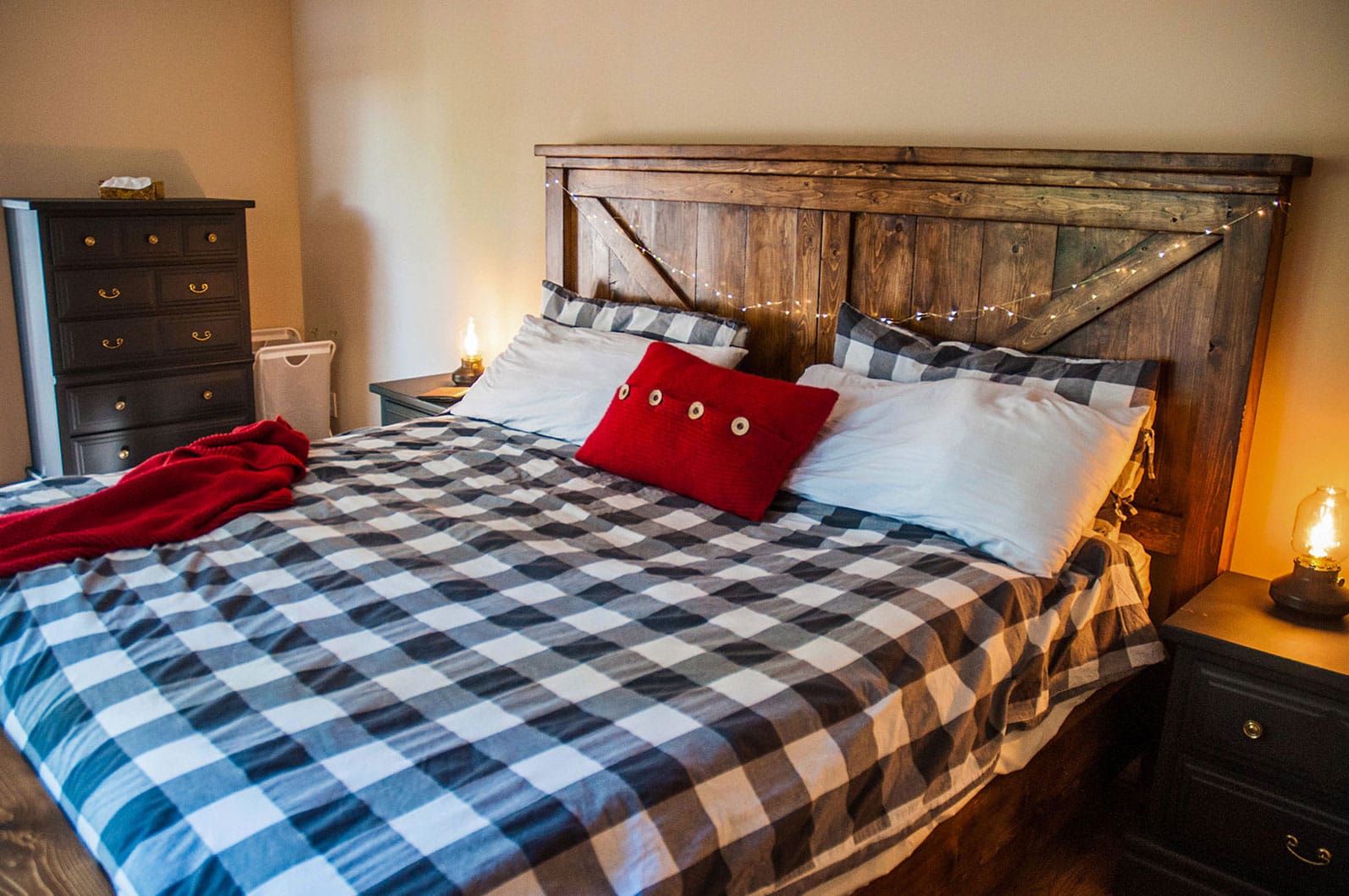 A cozy bedroom features a custom-made wooden king bed frame, accented by a bright red pillow. The rustic headboard and warm lighting from bedside lamps create an inviting and comfortable atmosphere.