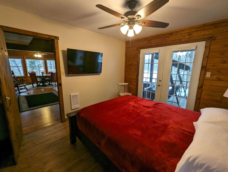 A cozy bedroom features a queen bed with a deep red blanket, a wall-mounted TV, and French doors leading to an outdoor space. A ceiling fan hangs above, and wooden walls add to the rustic charm.