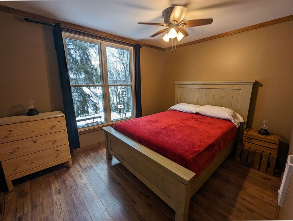 A loft bedroom features a queen bed with a rustic wooden frame. The room includes a large window offering views of the snowy outdoors, a ceiling fan, and natural wood furniture, creating a warm atmosphere.