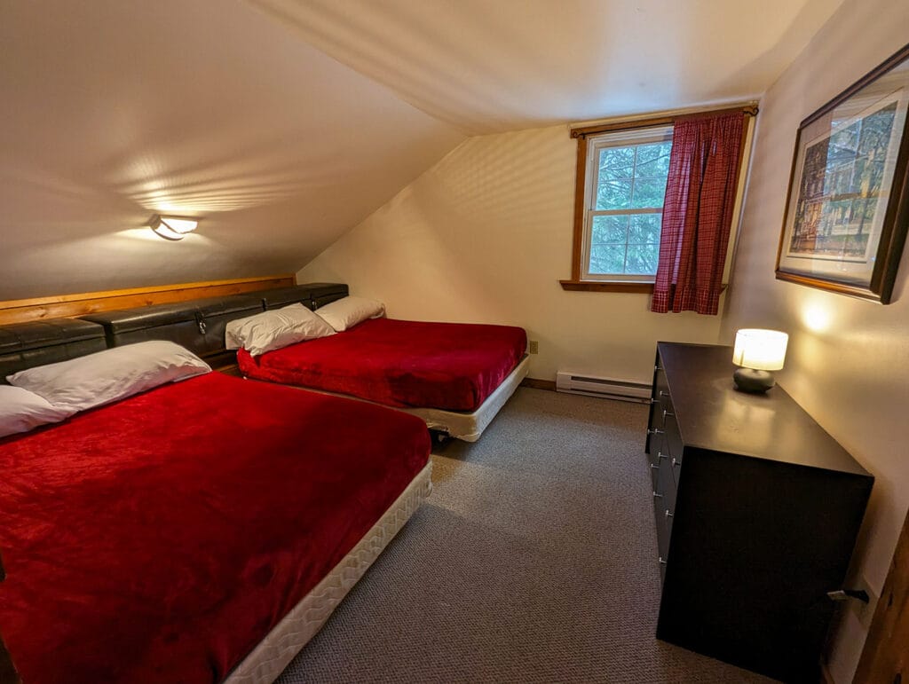 A cozy bedroom with a sloped ceiling features two double beds, each topped with a red blanket. The room offers excellent storage with a spacious dresser, bedside table, and a built-in headboard shelf.