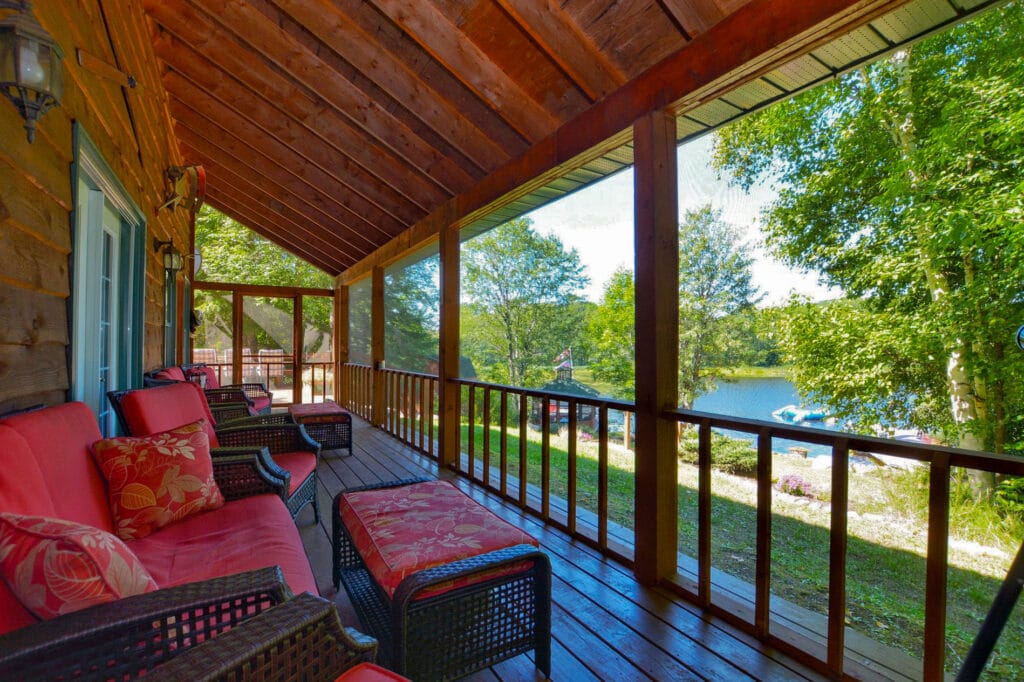 A spacious screened Muskoka room with wooden floors and ceiling features comfortable seating with red cushions. The room overlooks the lush greenery and lake, perfect for unwinding in a serene setting.
