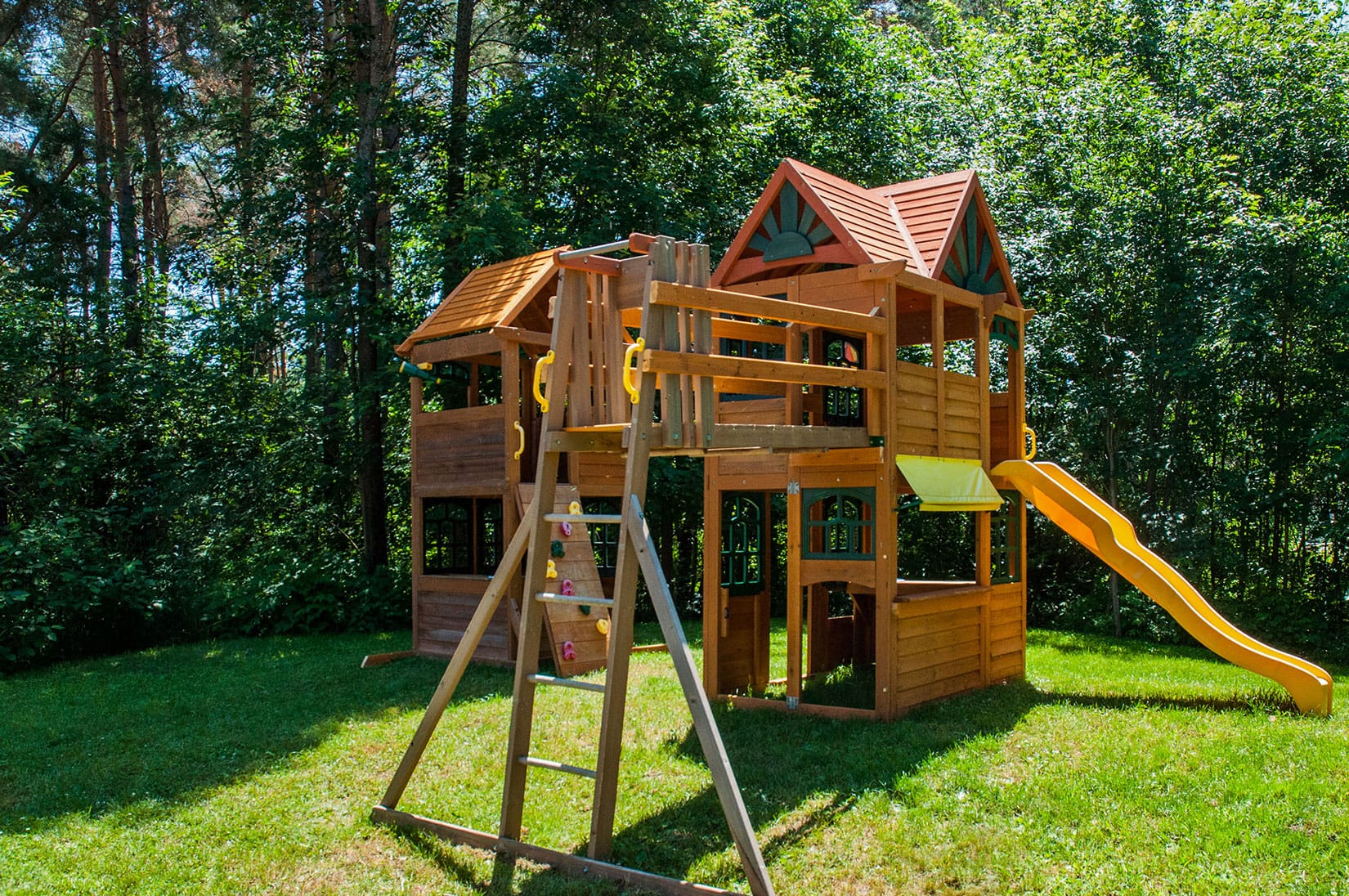 A spacious wooden playset stands in the front yard, featuring two levels with slides, ladders, and climbing walls. Surrounded by lush trees, it offers a fun and safe space for kids to play.