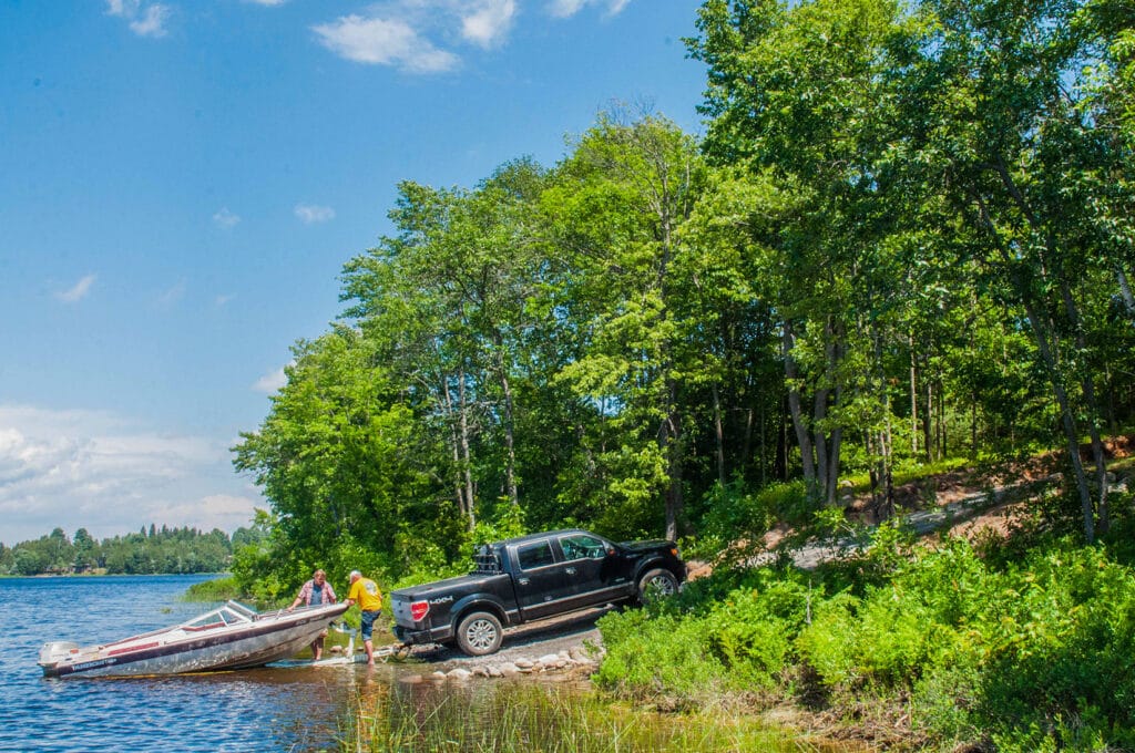 A truck with a boat trailer is backed into the private launch area at the lake, surrounded by lush greenery. Two people stand near the water, guiding the boat into the lake.