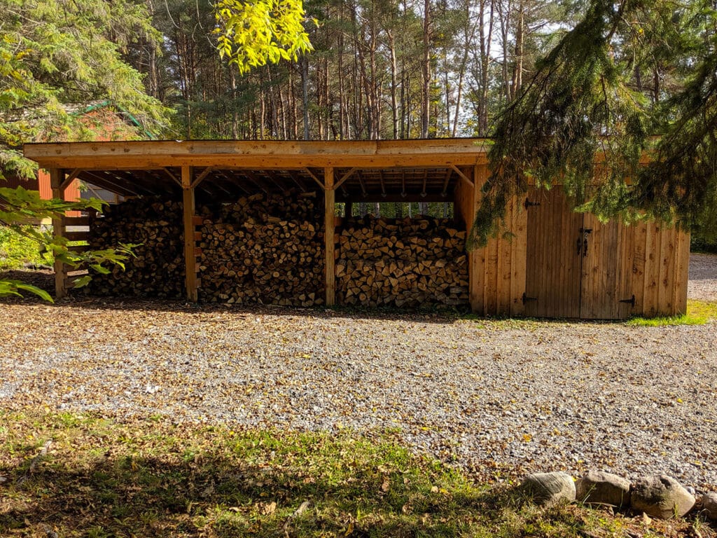 A sturdy wooden woodshed stocked with neatly stacked firewood, surrounded by tall trees and gravel paths, ensures you have ample supply for comfortable nights by the fire.