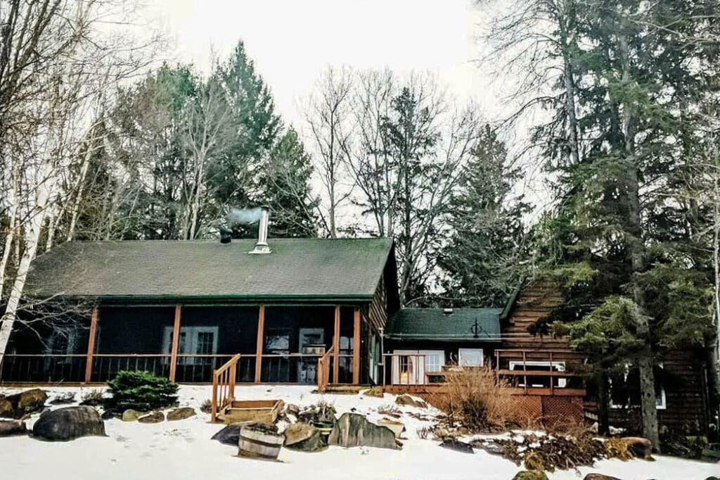 The cottage stands against a backdrop of tall, leafless trees, with snow covering the ground. Smoke rises gently from the chimney, hinting at cozy warmth inside the rustic log structure.