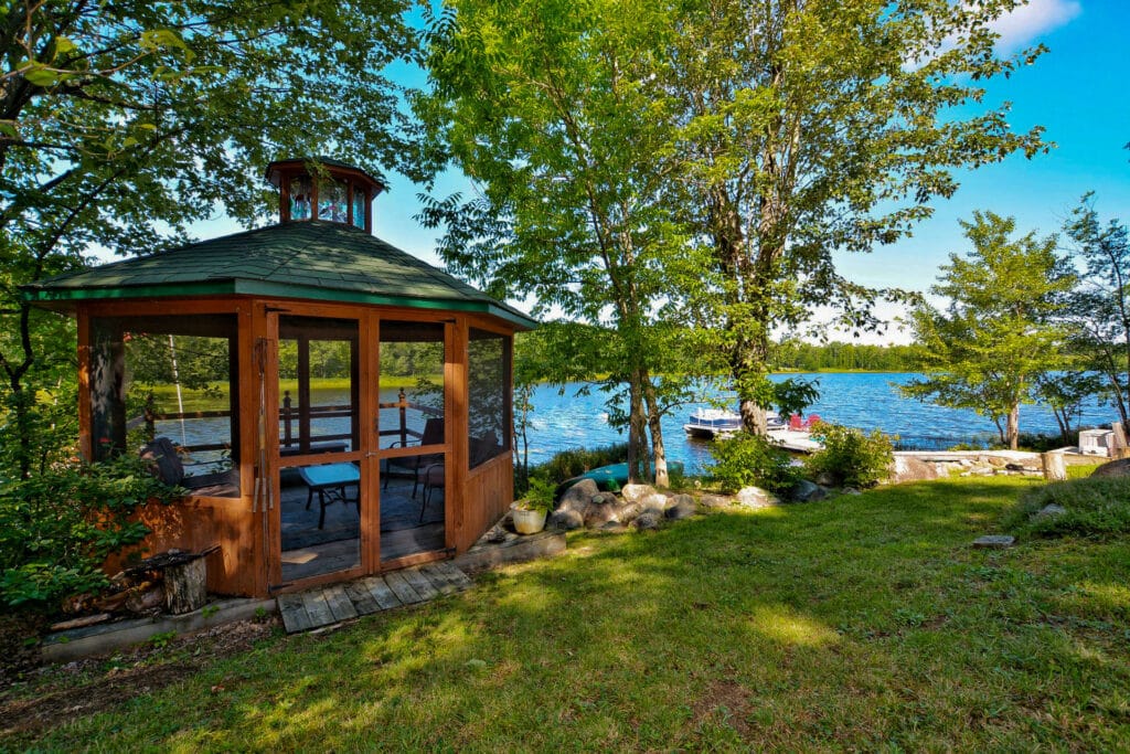 A screened gazebo sits among trees with a peaceful view of the lake. Inside, inviting seating offers a perfect spot to relax and enjoy the natural surroundings and gentle breeze.